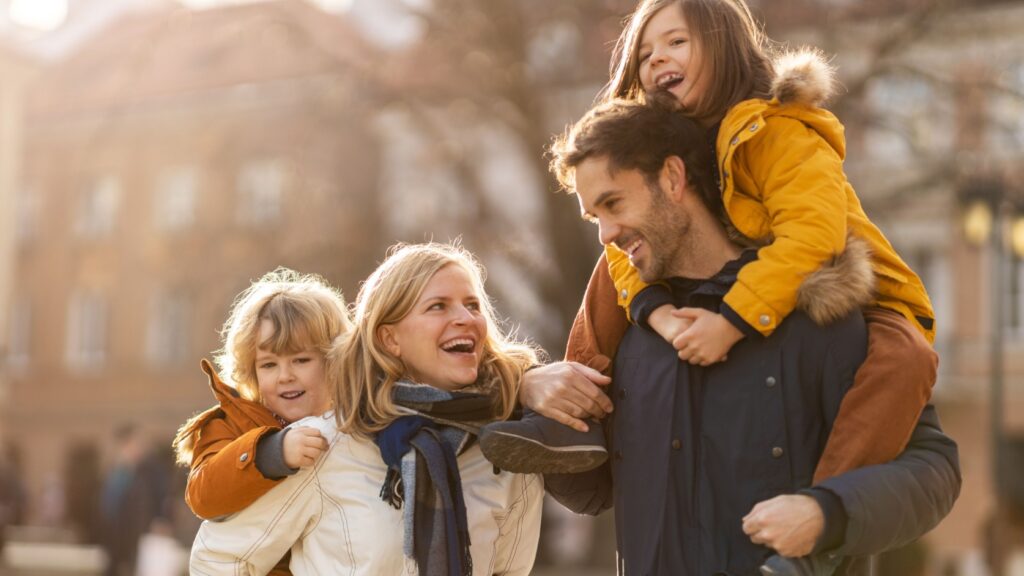 A husband and wife both give piggybacks to their young children in the middle of a city.