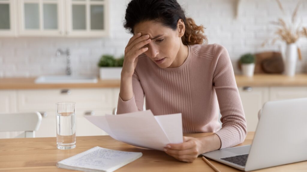 Stressed young woman looking at paperwork while using laptop.