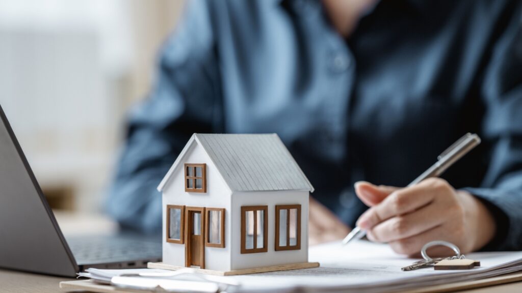 A person signing a document with a model house on the table.